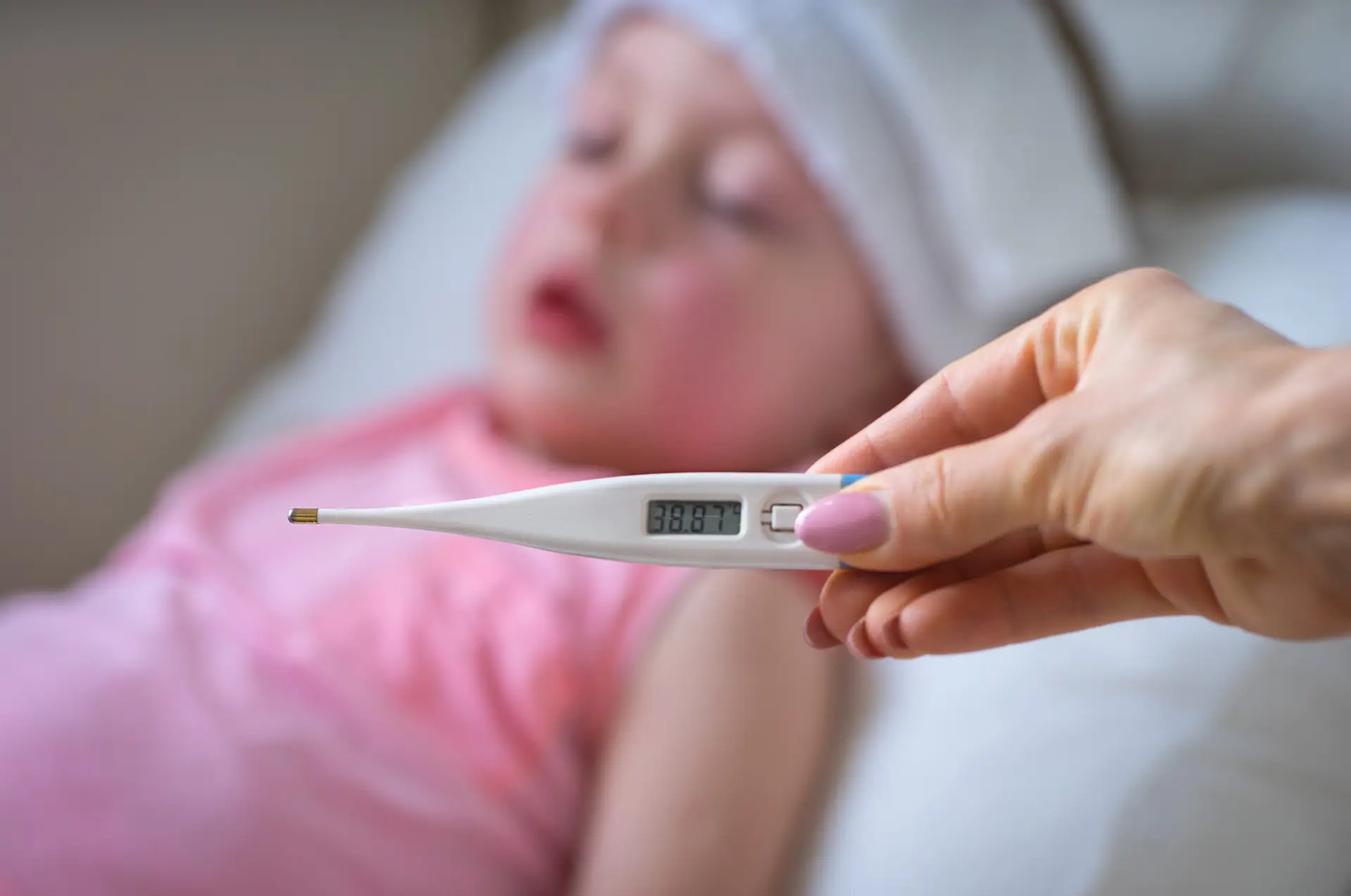 Sick child with high fever laying in bed and  holding thermometer.  Compress on forehead.